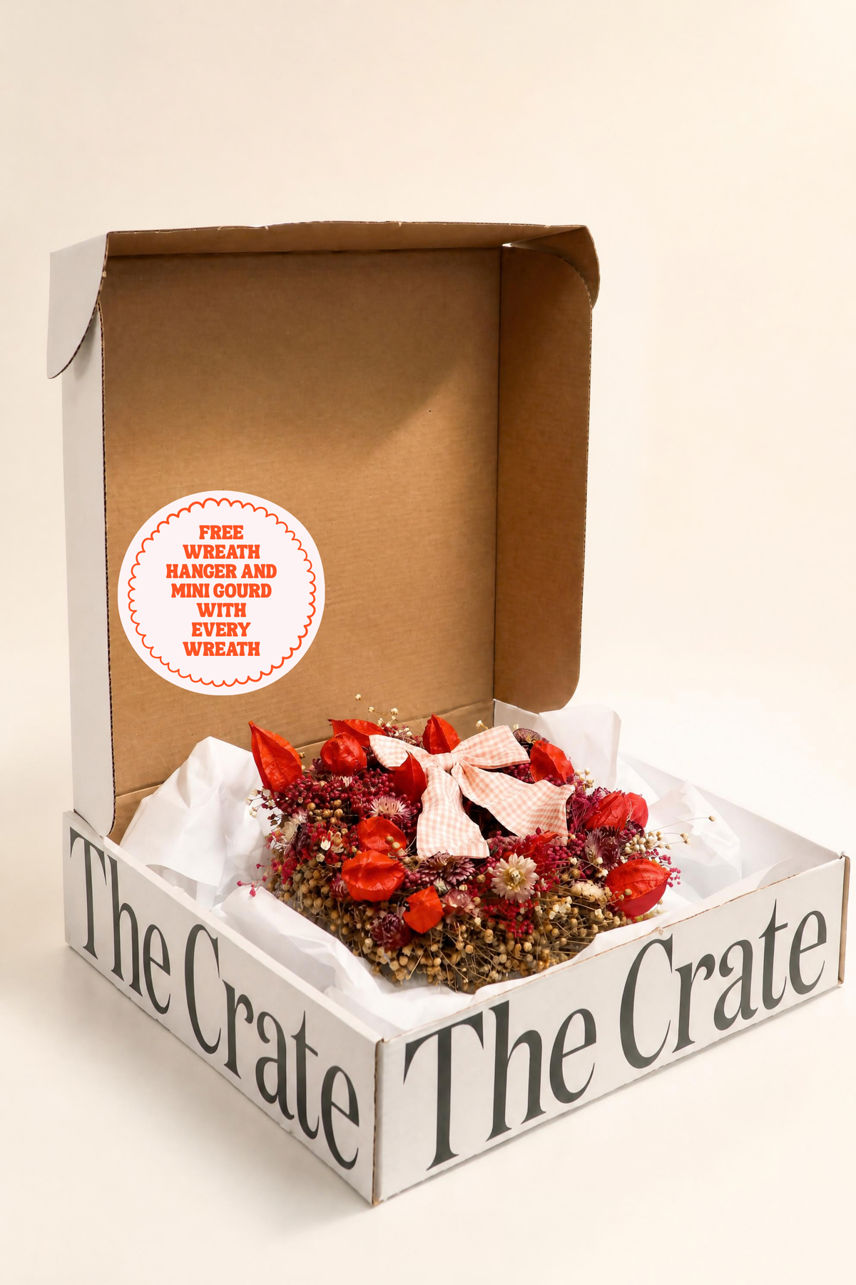 A floral arrangement gift box from The Crate, featuring a dried flower wreath with red berries, a pink bow, and small mini coeds nestled in white tissue paper inside an open cardboard box.