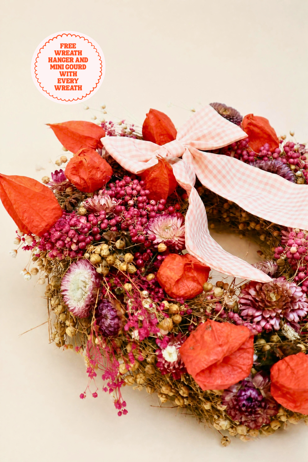 A dried floral wreath with orange-red paper lanterns, pink and purple flowers, and a large white gingham bow, photographed on a light beige background with a circular promotional badge in the top left.