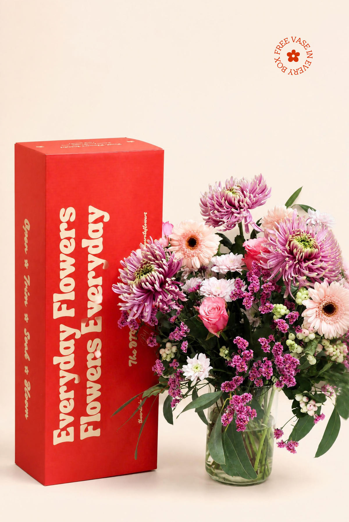 A vibrant red gift box with white text next to a colorful bouquet of pink, purple, and white flowers in a clear glass vase, set against a solid beige background.