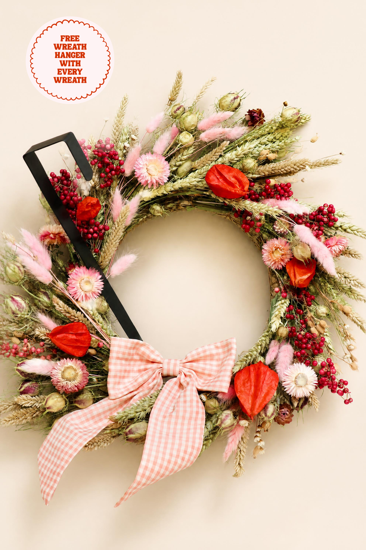 Flat lay close-up of a handcrafted dried flower wreath showing detailed pink strawflowers, fuzzy bunny tails, wheat stems, orange Chinese lanterns, and a pink gingham bow alongside a black metal over-door hanger.