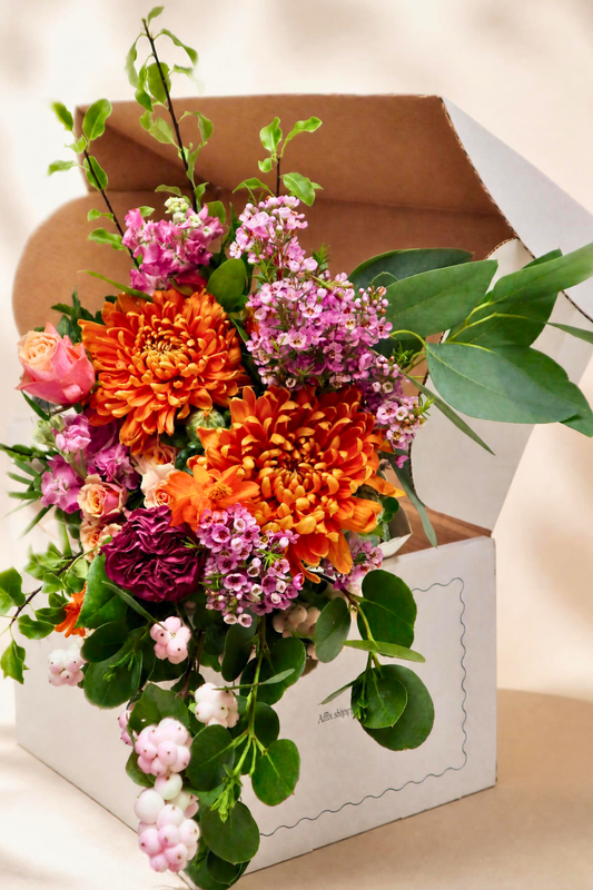 Vibrant close-up of a seasonal flower bouquet overflowing from a white presentation box, showcasing orange chrysanthemums, pink roses, purple carnations, soft pink berries, and fresh eucalyptus foliage. Perfect luxury floral gift arrangement for birthdays, anniversaries, or special occasions.