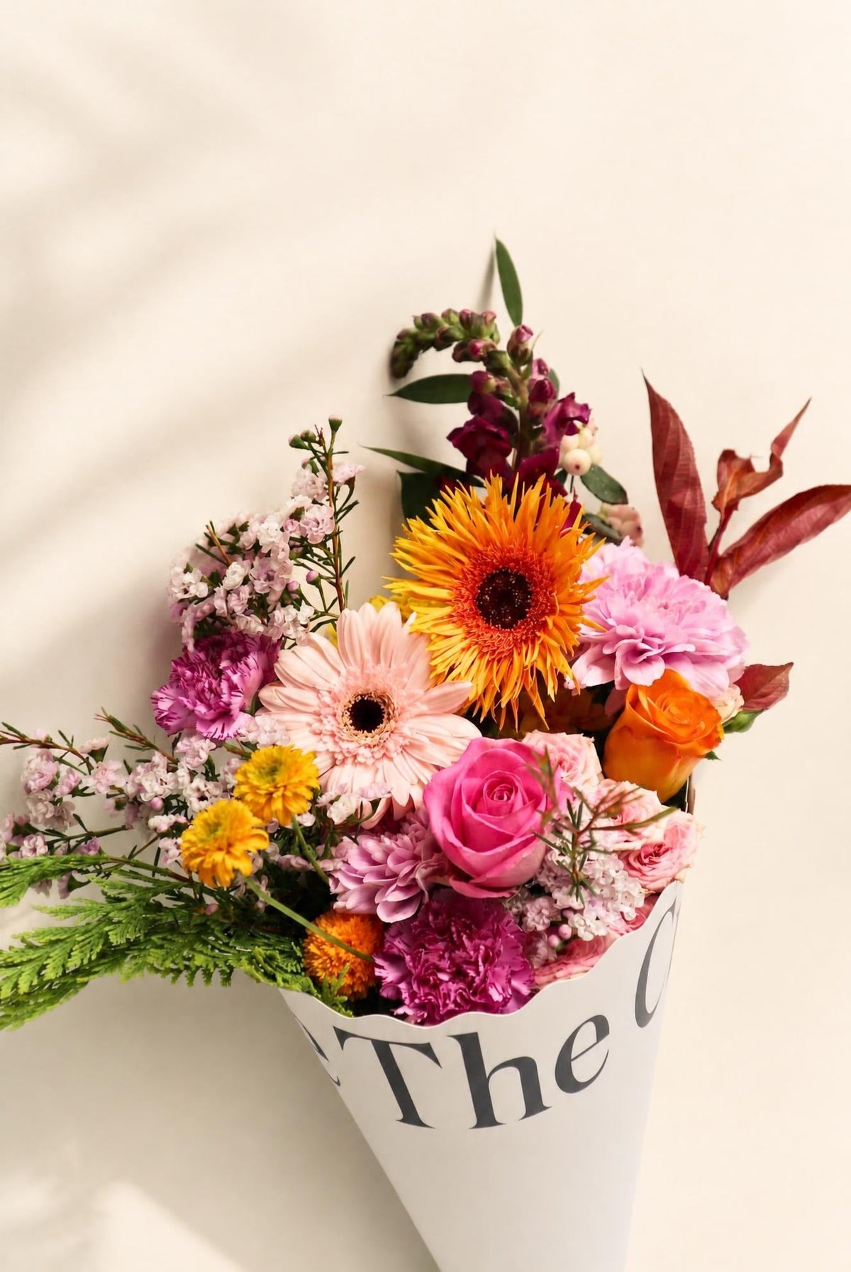 The Crate's Weekly Bunch: A vibrant mixed floral bouquet featuring pink roses, peach and orange gerbera daisies, purple blooms, and lush greenery, elegantly wrapped in signature white paper for fresh flower delivery.