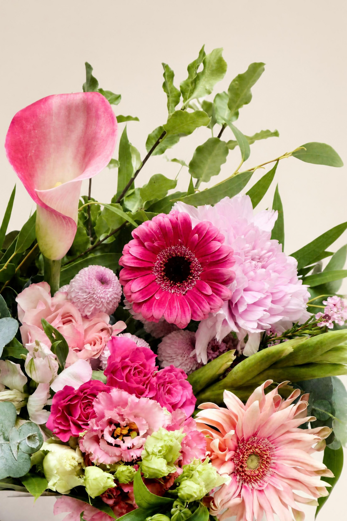 A vibrant floral bouquet featuring pink and magenta flowers, including a calla lily, gerbera daisies, roses, carnations, and chrysanthemums, accented with green foliage, set against a plain light beige background.