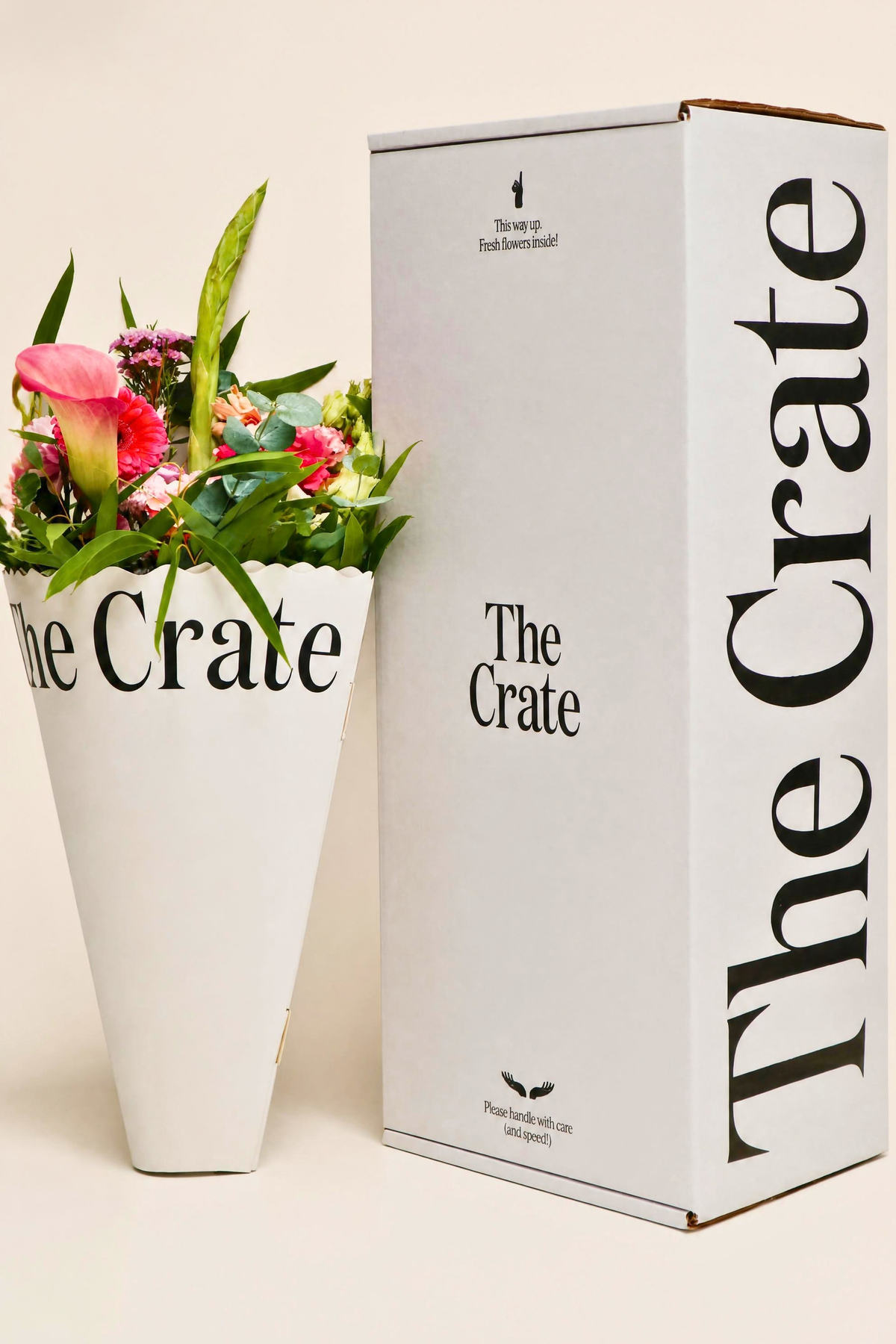 A white cardboard box printed with 'The Crate' stands beside a white vase filled with pink and red flowers and green leaves, also printed with 'The Crate,' on a light beige background.