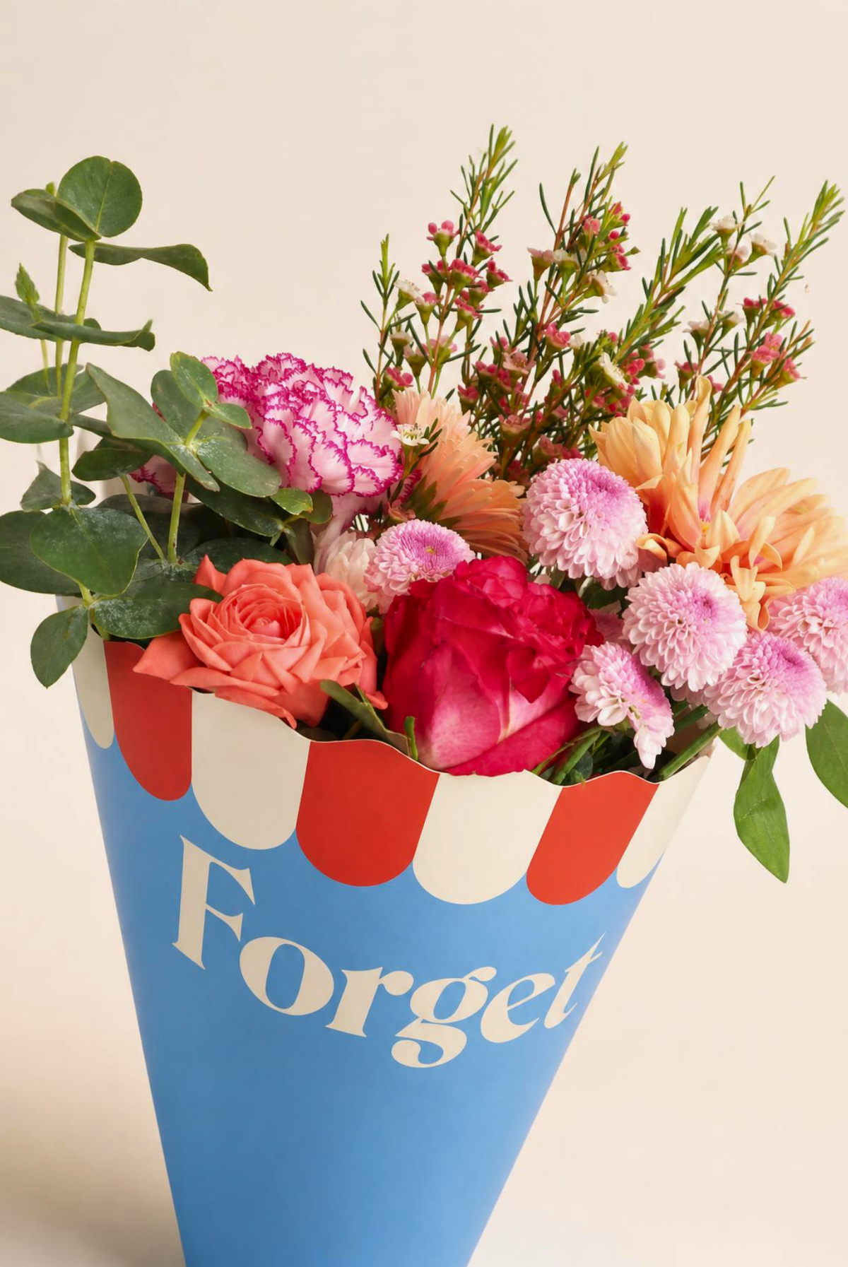 A floral arrangement in a blue cone-shaped container with red and white scalloped trim and the word 'Forget' printed on it, set against a plain light beige background.