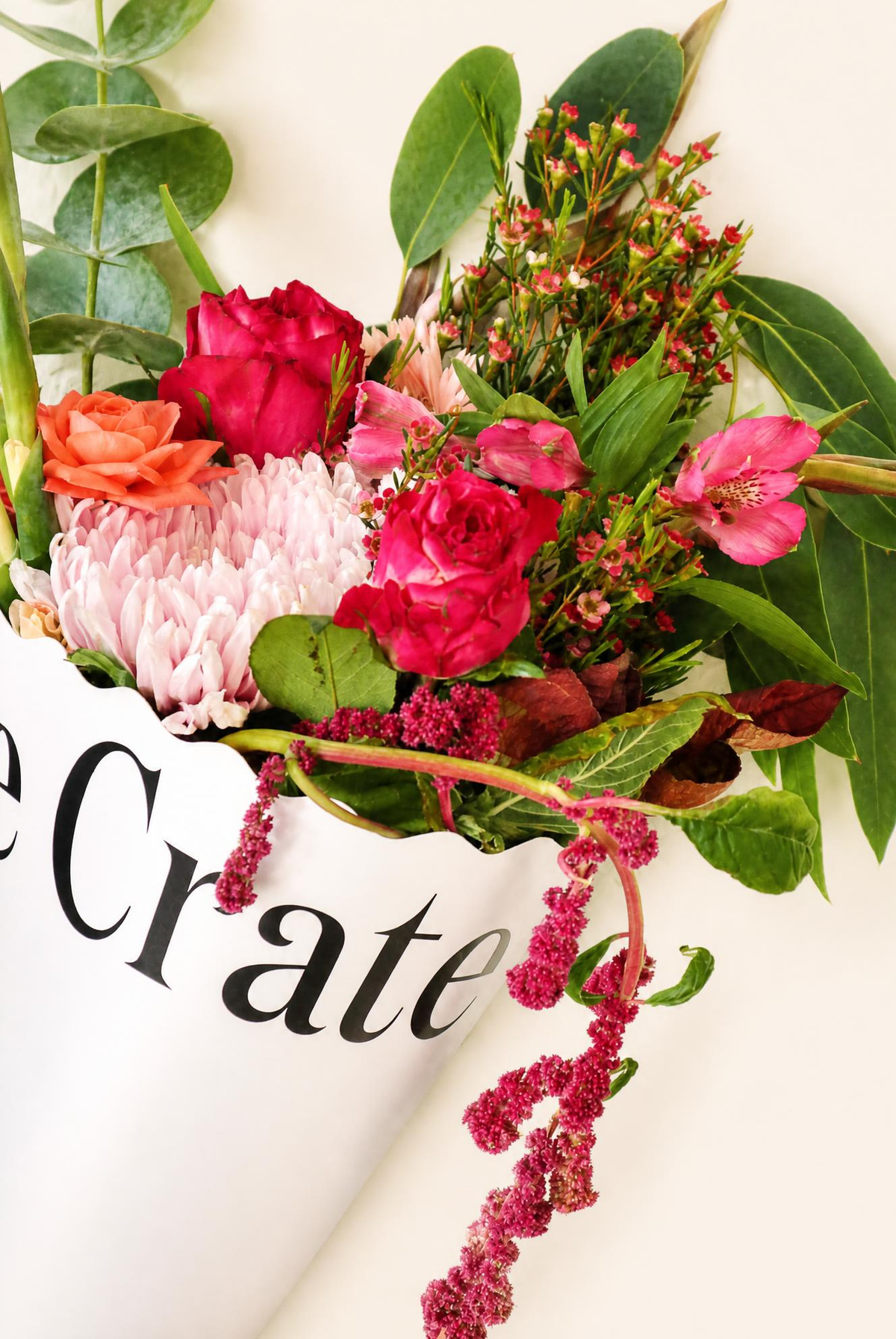 A close-up of a floral arrangement in a white container with black text reading 'Crate', featuring bright pink and red flowers, green leaves, and burgundy amaranth stems against a light cream background.