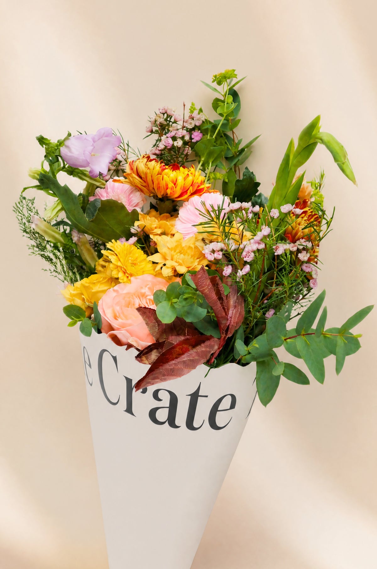 A colorful floral arrangement in a white cone-shaped box printed with the word 'Crate', set against a soft beige background.