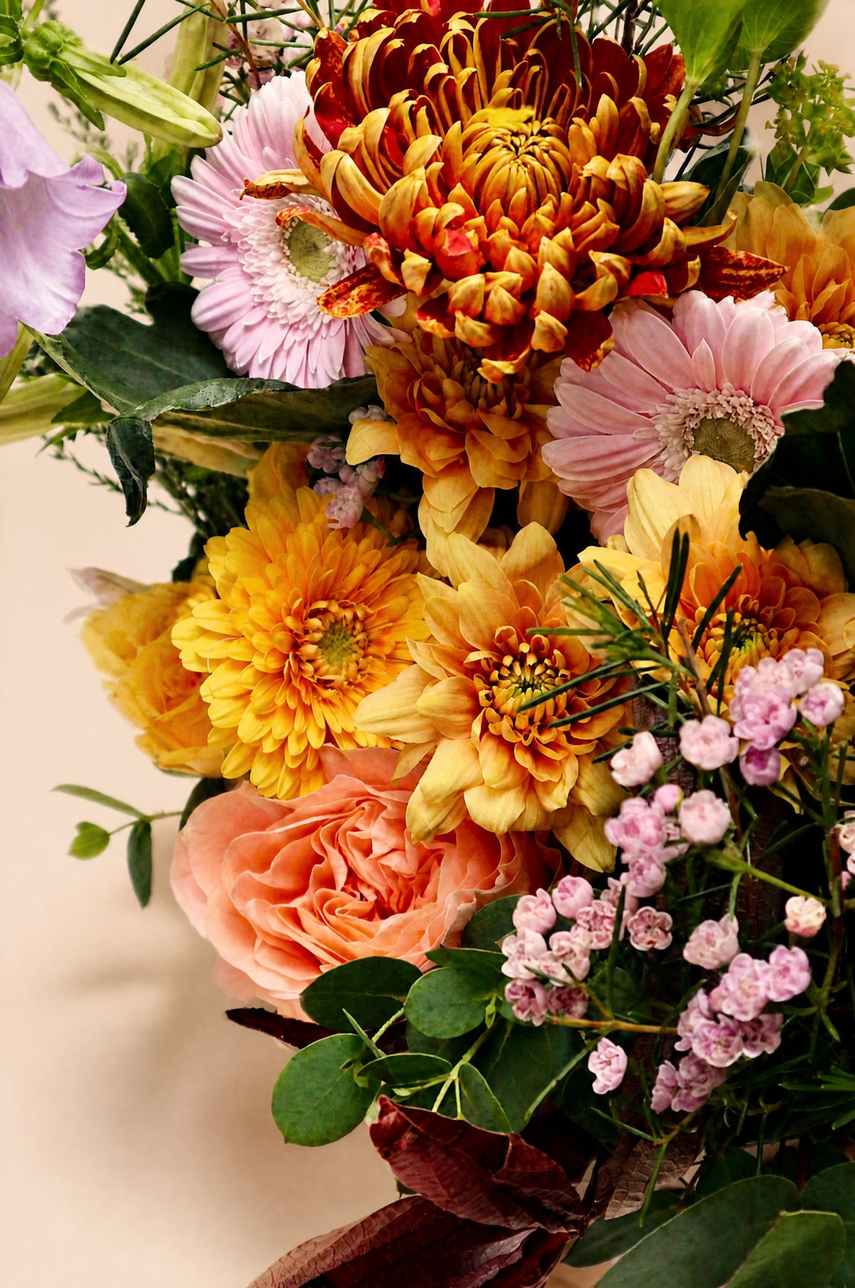 A close-up of a vibrant floral arrangement featuring yellow and orange chrysanthemums, pink daisies, peach roses, and small pink baby's breath flowers with green foliage, set against a plain beige background.