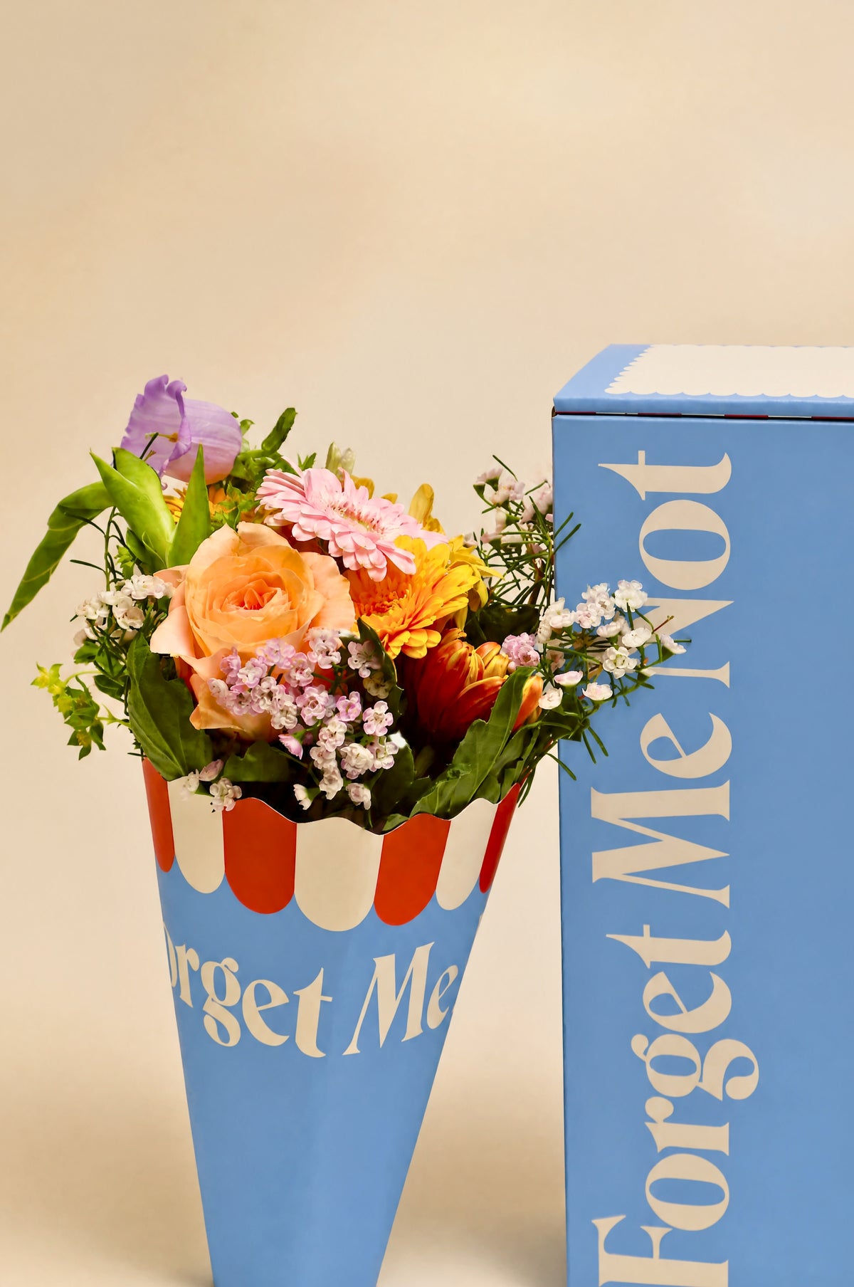 A bouquet of multicolored flowers arranged in a blue cone-shaped container with red and white stripes, placed beside a tall blue box printed with the words 'Forget Me Not.'.