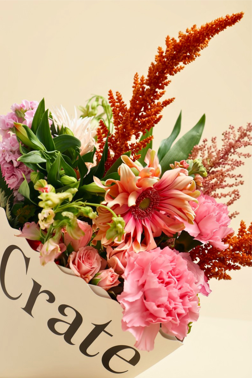 A vibrant floral arrangement in a white paper box printed with the word 'Crate', featuring pink carnations, orange astilbe, and green foliage on a light beige background.