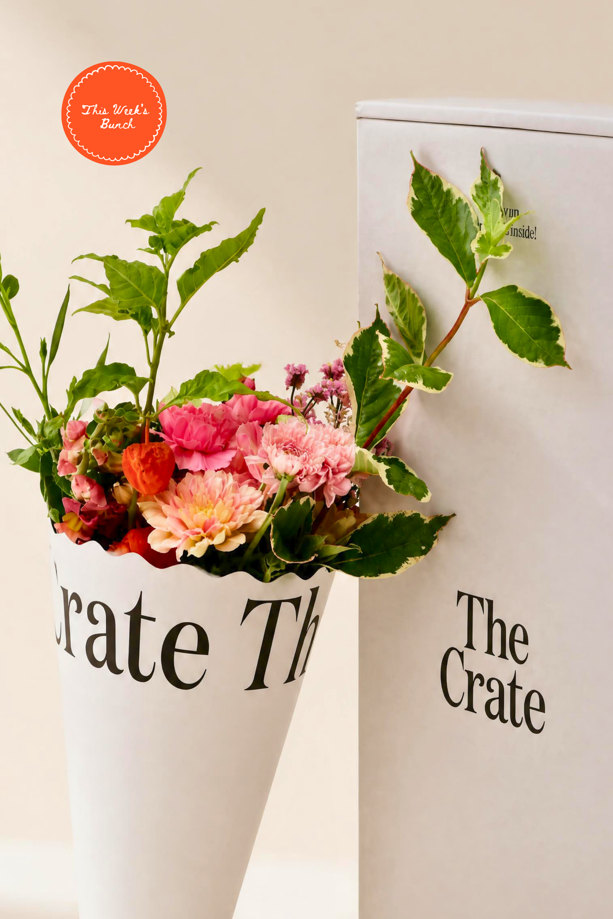 Fresh pink dahlias, carnations, and peach flowers in a white branded paper cone next to a signature white delivery gift box by The Crate.