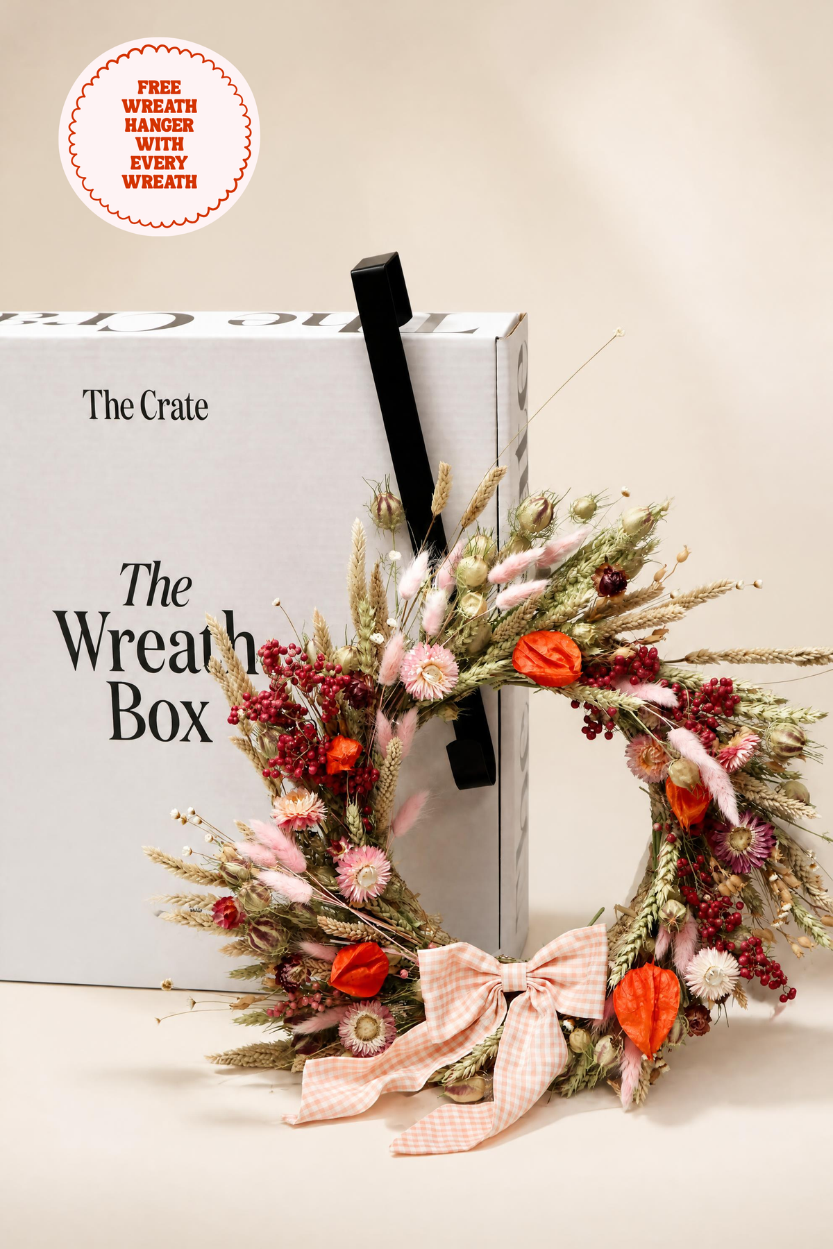 The Crate dried flower wreath resting against "The Wreath Box" gift packaging with a black door hanger hook. Styled with pink strawflowers, orange physalis, wheat, and a pink gingham ribbon.