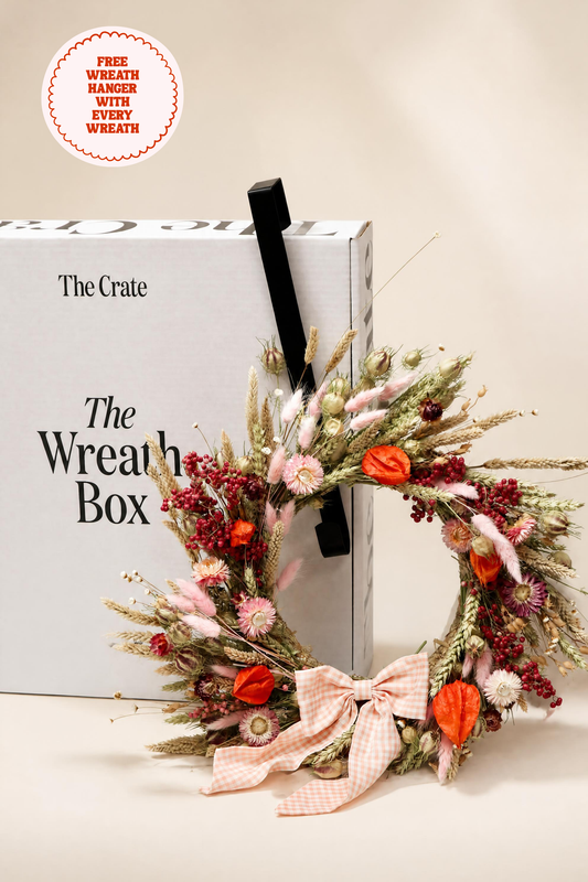 The Crate dried flower wreath resting against "The Wreath Box" gift packaging with a black door hanger hook. Styled with pink strawflowers, orange physalis, wheat, and a pink gingham ribbon.