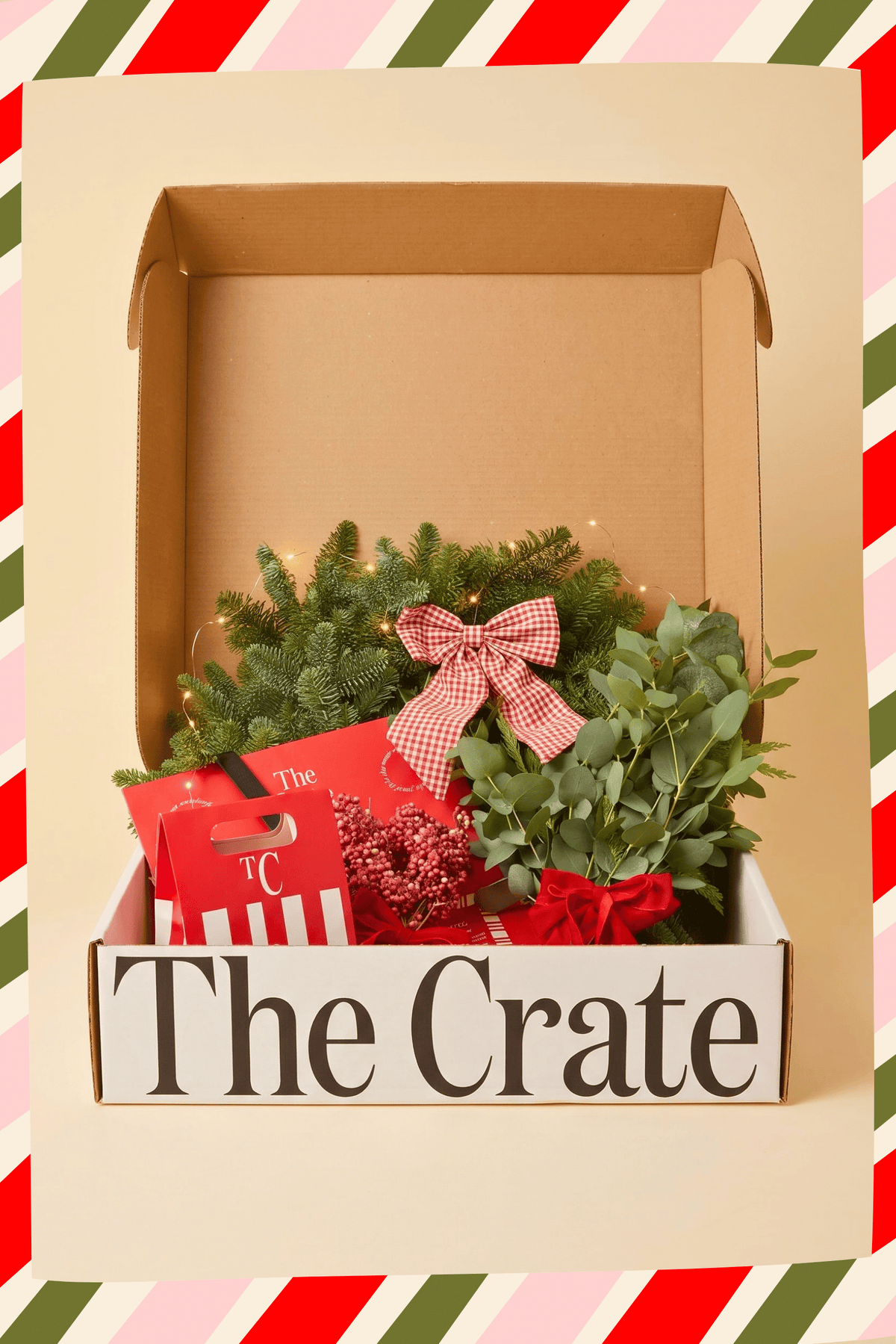 A Christmas-themed DIY Wreath gift box from The Crate, featuring green pine branches, eucalyptus, red berries, and a pink gingham bow inside a white wooden box labeled 'The Crate'.