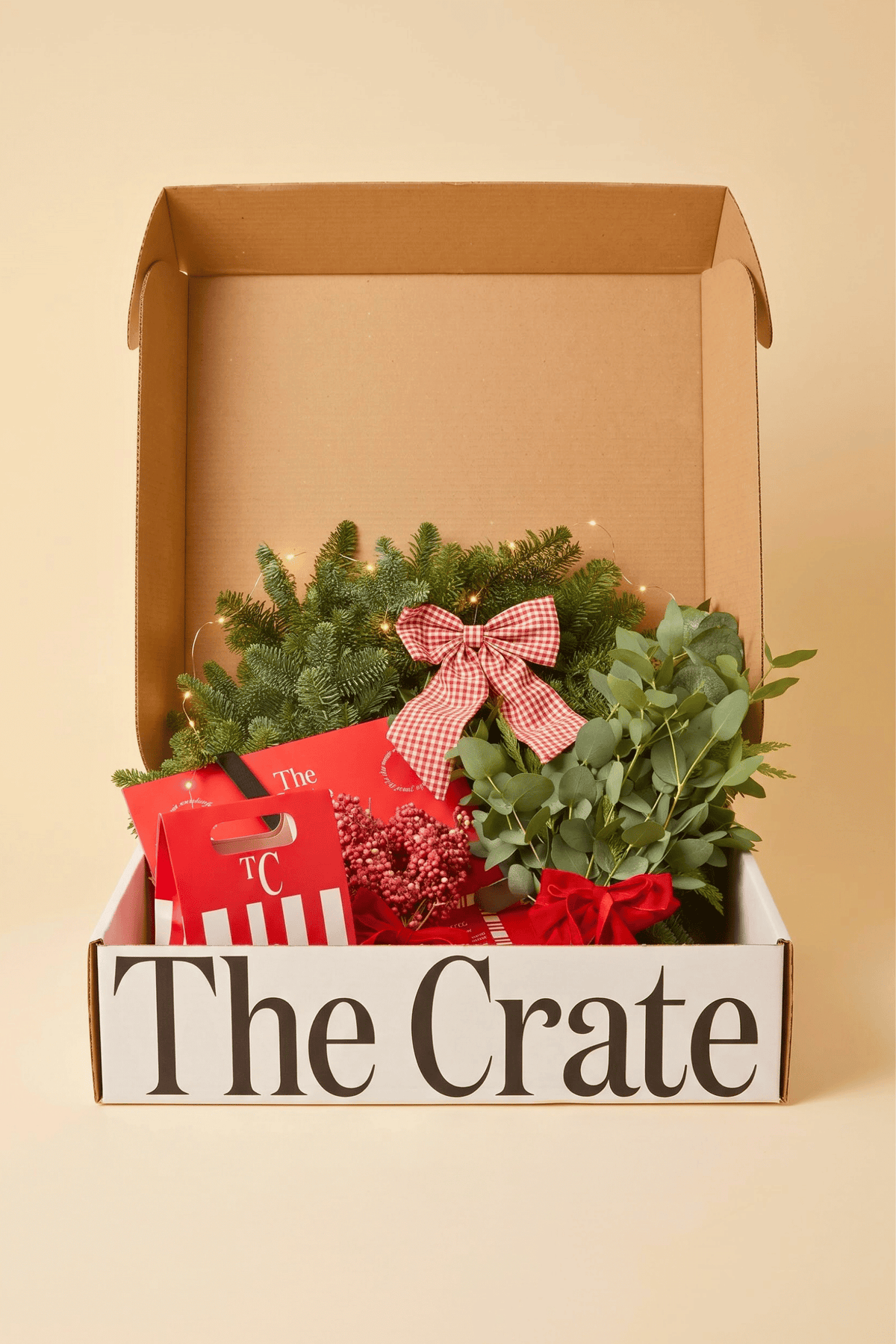The Crate's DIY Christmas Wreath Kit gift box labeled 'The Crate' filled with holiday greenery, red ribbon, and red gift packaging on a warm beige background.