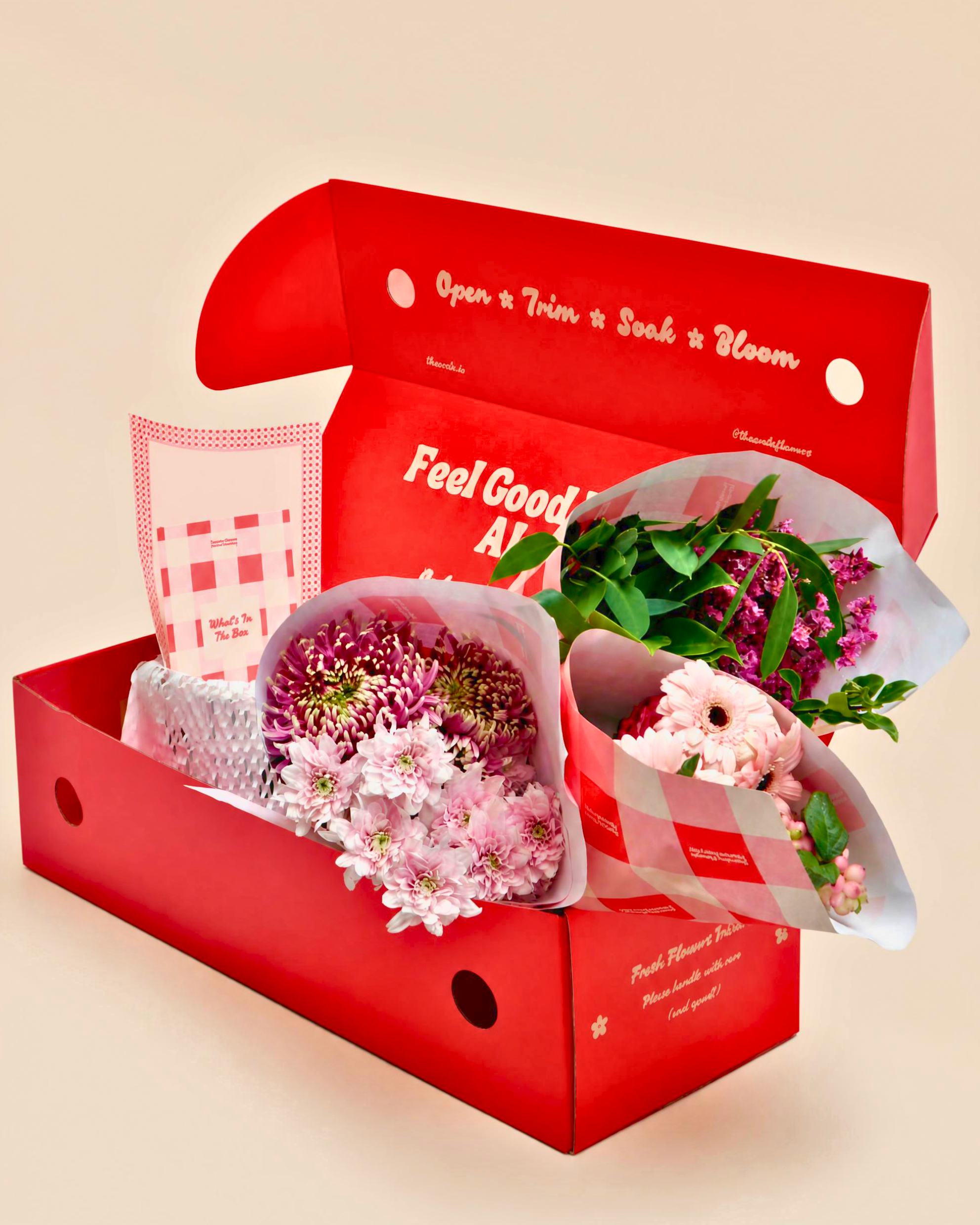A red gift box filled with pink and white flower bouquets, a small card, and a wrapped item, photographed on a plain warm beige background. The box lid has printed text including 'Open & Trim & Suck & Bloom' and 'Feel Good All'.
