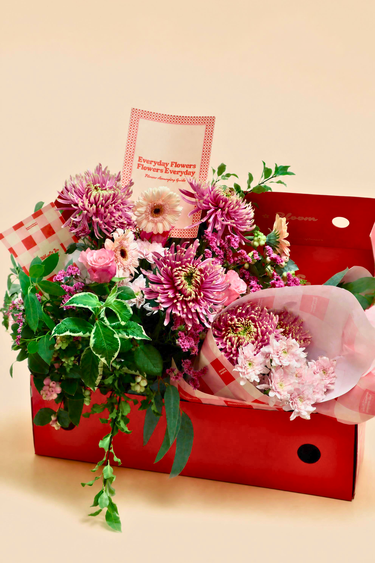 A vibrant flower bouquet arranged inside a red gift box, featuring pink chrysanthemums, green foliage, and a small card that reads 'Everyday Flowers Flowers Everyday'.