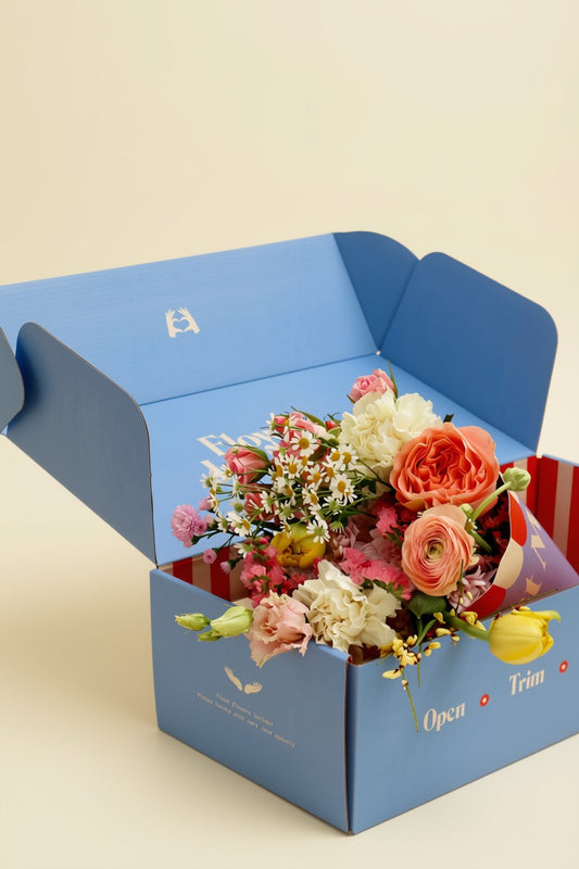 A blue flower cardboard gift box with the words 'Open Trim' printed on the front, shown open and filled with a colourful bouquet of fresh flowers including pink, peach, white, and yellow blooms, set against a solid light beige background.