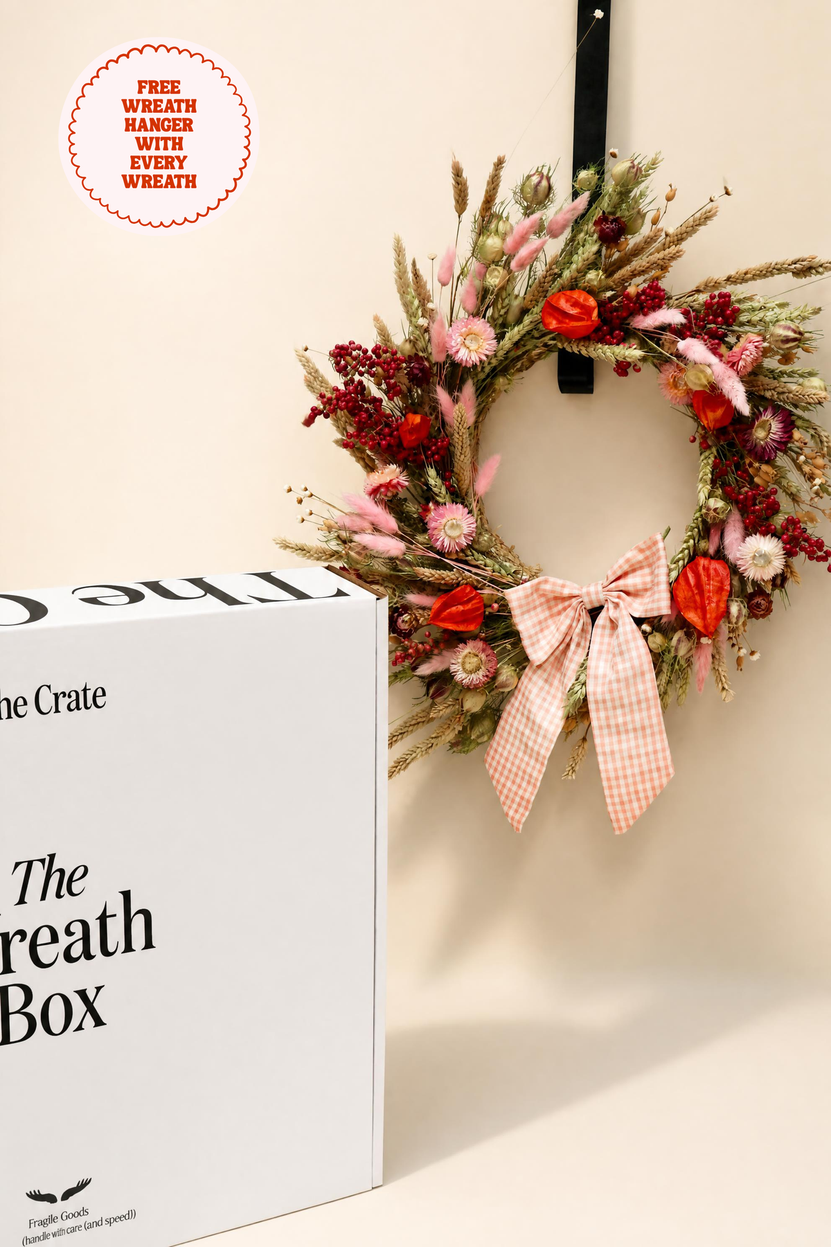 The Crate "The Wreath Box" eco-friendly packaging displayed next to a pink and orange dried flower wreath with a gingham bow hanging on a black metal hanger. Includes free wreath hanger offer.